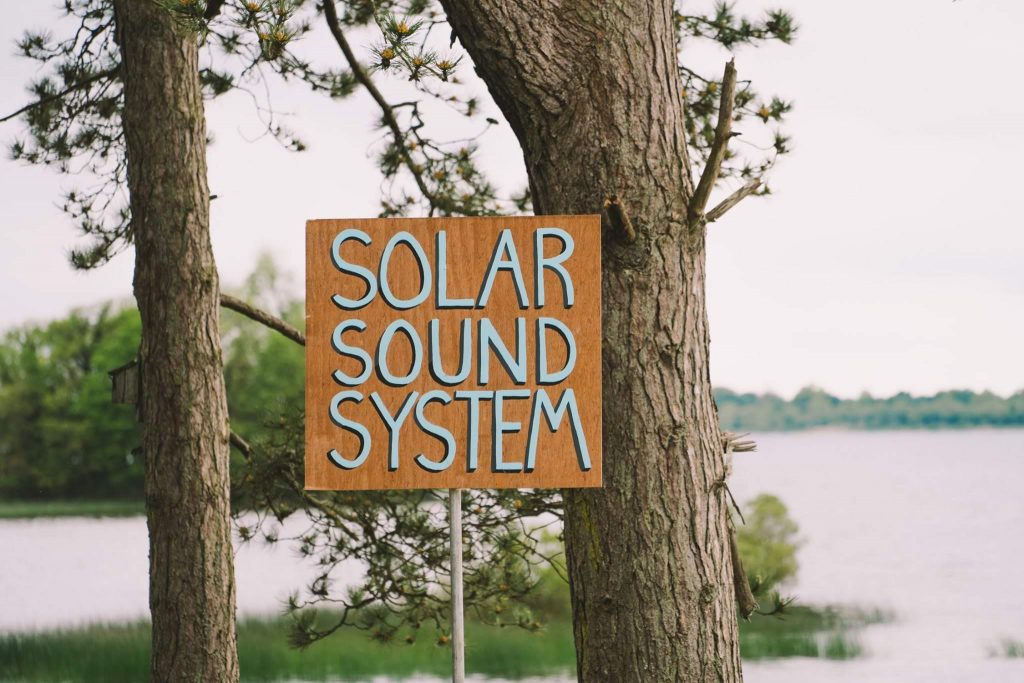 Wooden sign with the words 'Solar Sound System' in front of large trees and a lake